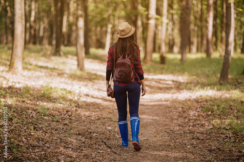 Stylish hipster young woman walking in the forest, carrying a backpack in the forest on sunset light in the spring season, looking at amazing woods, travel concept.