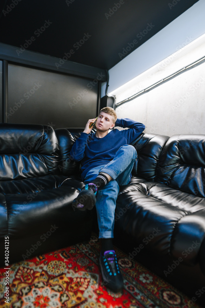 Stylish young man sitting on a black leather sofa and talking on the phone. Trendy guy in jeans and trendy sneakers makes a phone call and relaxes on the sofa indoors. Vertical