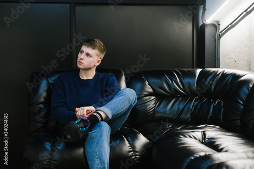 Young man in casual clothes sits on a leather sofa in a dark room, looking away with a serious face. Copy space. The guy is resting on a leather sofa.