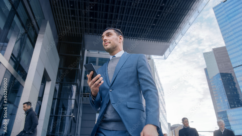 Caucasian Businessman in a Suit is Using a Smartphone on a Street in Downtown. Other Office People Walk Past. He Smiles and Looks Successful. He's Browsing the Web on his Device. Caucasian Businessman in a Suit is Using a Smartphone on a Street in Downtown. Other Office People Walk Past. He Smiles and Looks Successful. He's Browsing the Web on his Device.