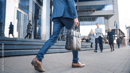 Close Up Leg Shot of a Businessman in a Suit Commuting to the Office on Foot. He's Carrying a Leather Case. Other Managers and Business People Walk Nearby. Cloudy Day on a Downtown Street.