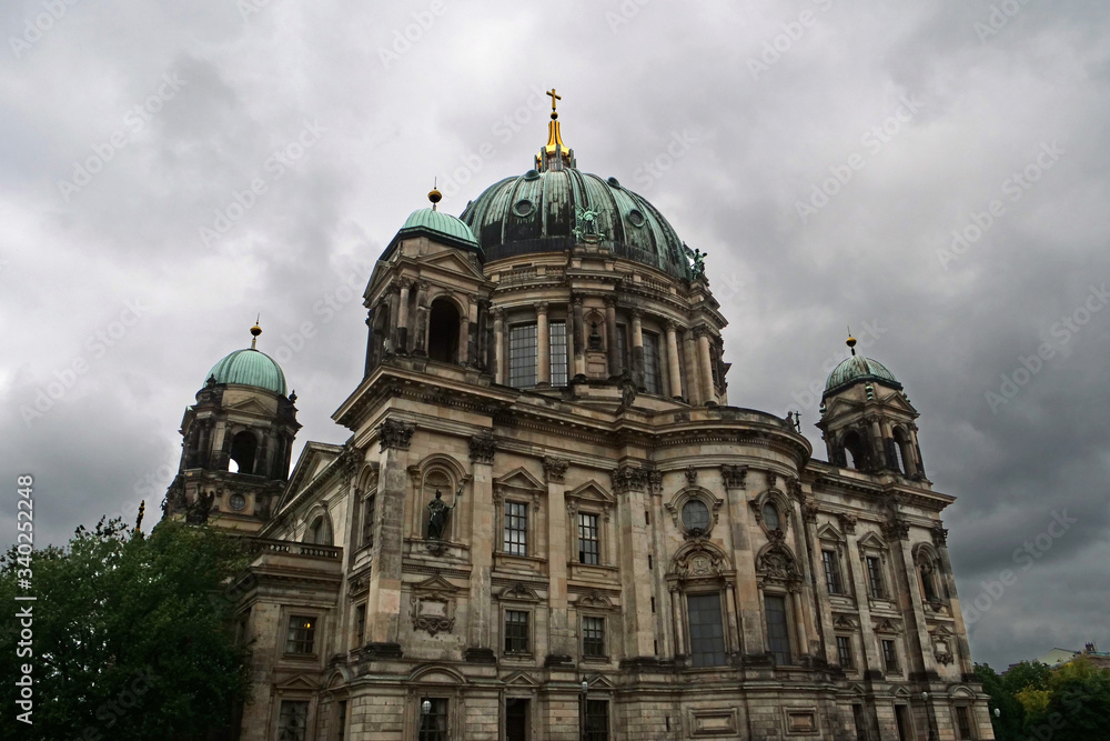 View of the upper part of the cupola of the Berlin Cathedral with the turquoise copper roof and golden cross on top in front of an impressive sky with white grey clouds; Berlin, Germany, Europe
