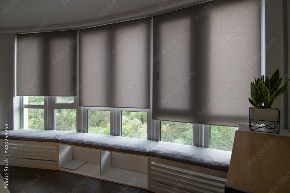 Motorized roller shades in the interior. Automatic roller blinds beige  color on big glass windows. Home luxury curtaines are above the windosill  with pillows. Summer. Green trees outside. Photos | Adobe Stock
