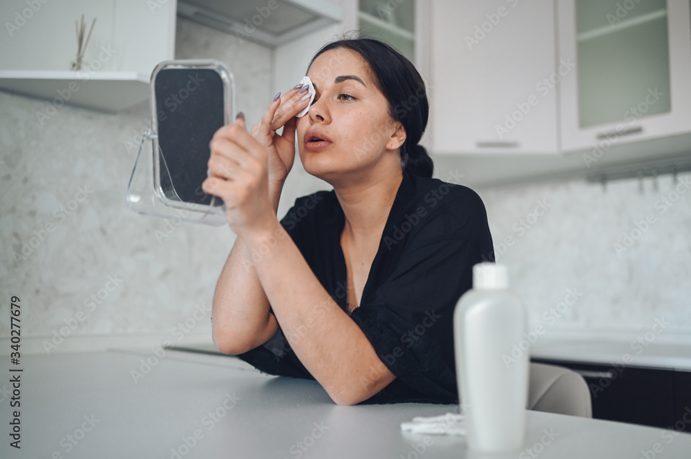 Obraz premium Beautiful brunette woman removing makeup from her face with mirror. Cute beautiful natural beauty girl cleaning face with cotton sponge. Cosmetology and spa, problem skin care, acne treatment concept