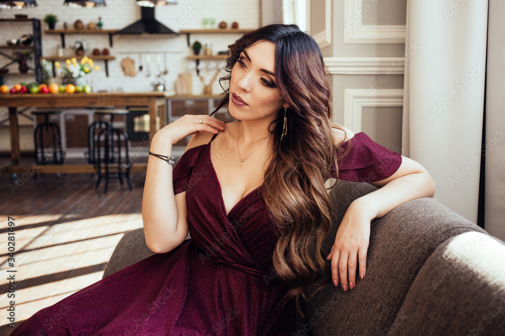 A portrait of young beautiful woman in vinous dress sitting on the sofa at home. Sunny day. A portrait of young beautiful woman in vinous dress sitting on the sofa at home. Sunny day.