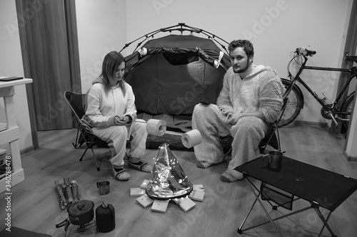 Couple sitting in folding chairs by the tent and bonfire. Camping in the apartment. Adventures at home, traveling around the room. Black and white. Funny, fried marshmallows, toilet paper