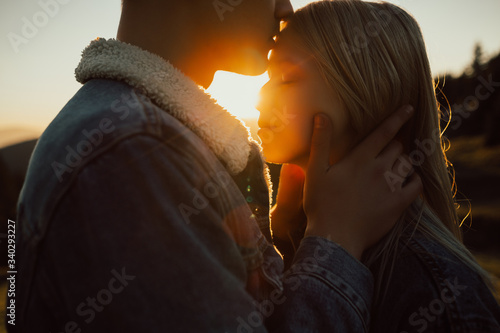 Cute young beautiful couple posing on mountain at sunset. Boyfriend kissing his girlfriend softly on the forehead, hipster, outdoor portrait, close up portrait. Cropped photo.