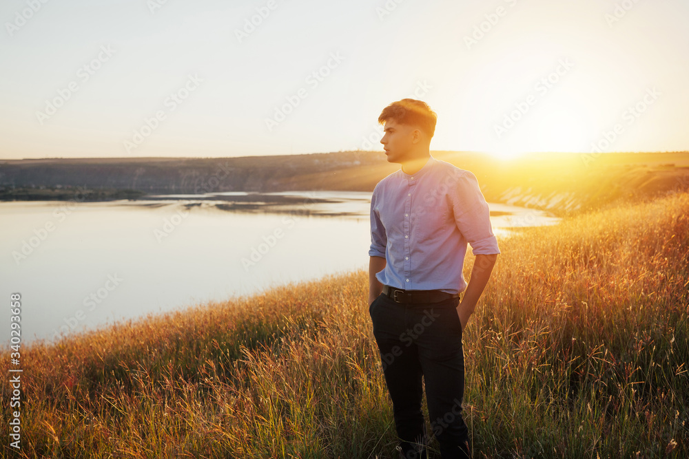 Free man in stylish classical clothes on sunset admire nature view of summer lake. Success. Travel. Good life.  Handsome man standing with hands on hips on hill and looking at lake. Copy space.