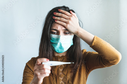 Sick young woman wearing a surgical mask and holding a digital thermometer that indicates she has over 38 degrees fever. Sick and worried female with fever and illness during pandemic.  photo