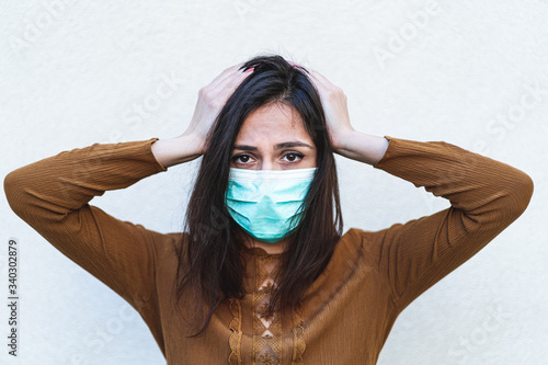 Disturbed annoyed young woman holding hands above head wearing a surgical mask feeling sick and worried about epidemic outbreak. Anxious lady touching head with stress migraine photo