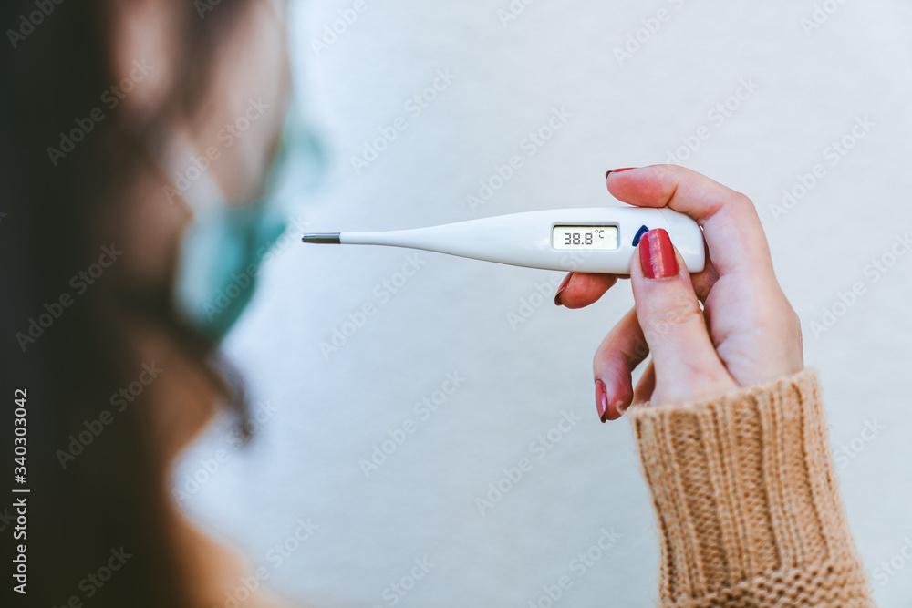 Φωτογραφία Close up back view of a sick young woman wearing a surgical mask and holding a digital thermometer that indicates she has over 38 degrees fever