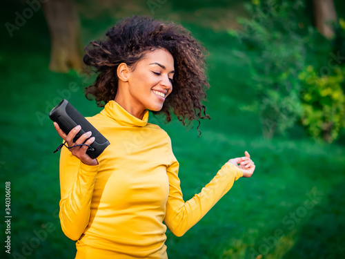Modern trendy african american girl listening to music by wireless portable speaker.Young beautiful woman enjoying, dancing in park.