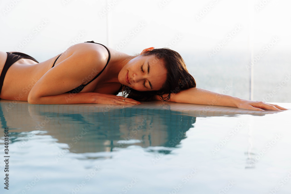Obraz premium Close up portrait of a beautiful happy woman resting on her hands at the side of a sun bathed swimming pool