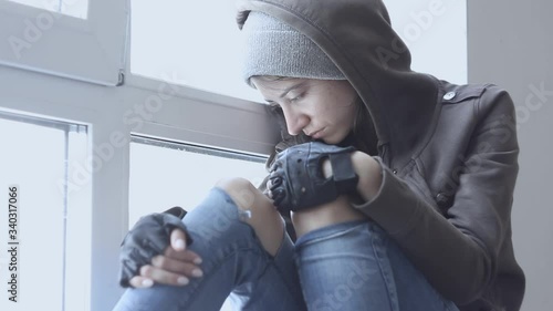 Lonely, depressed, upset young woman in hood is sitting on windowsill and crying in abandoned building. Homeless teenage girl ran away from home because of problems. Alcohol abuse, addiction concept.