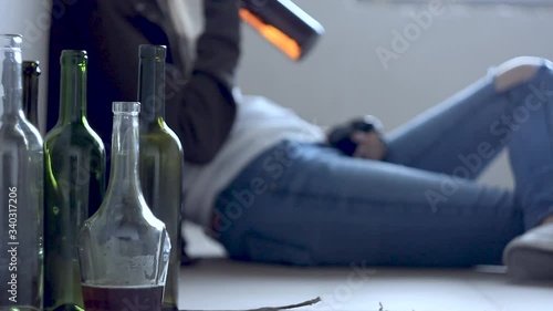 Closeup empty glass bottles. On background homeless drunk woman is sitting on cardboard on floor in abandoned house. Addicted teenage girl drunkard is drinking beer. Alcoholism, alcohol abuse concept.