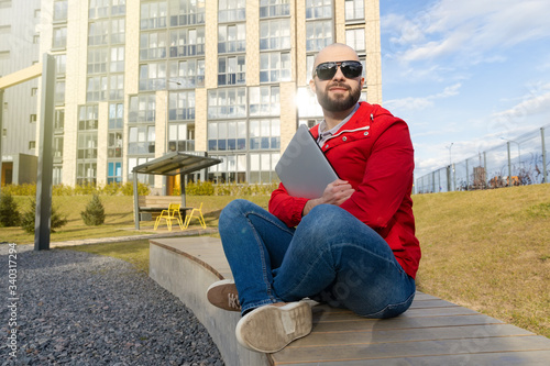 Portrait of a guy with a beard in a red jacket and jeans holding a laptop against the background of a city block. The concept of freelance work