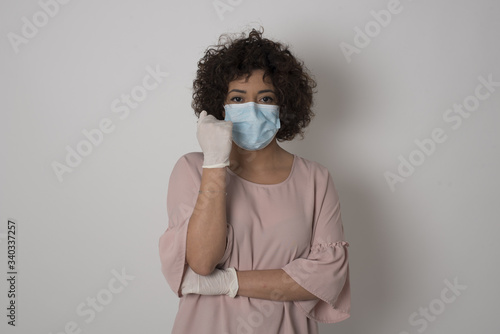 Isolated portrait of stylish European woman wearing medical mask with hand near face and looking to the camera with doubtful and skeptical expression suspect and doubt.  © Jihan