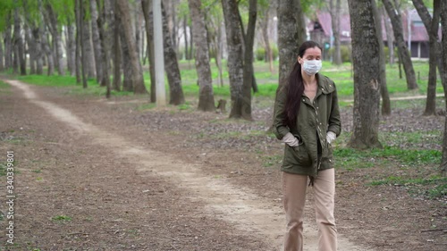 Wonderful cute Russian woman in protective medical face mask with long brown hair walks alone in summer forest, enjoys nature and fresh air, looks up on the sun at the sky outdoor. Isolation concept. photo