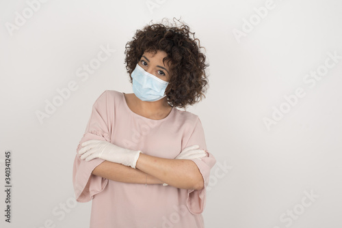 Waist up shot of beautiful self confident cheerful young woman wearing medical mask, crosses arms, happy to meet with colleague, dressed in fashionable clothes. © Jihan