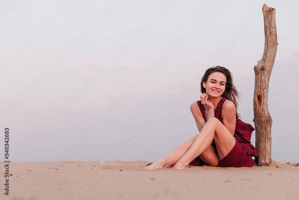 Girl sitting in the desert in the wind in a red dress near an old tree