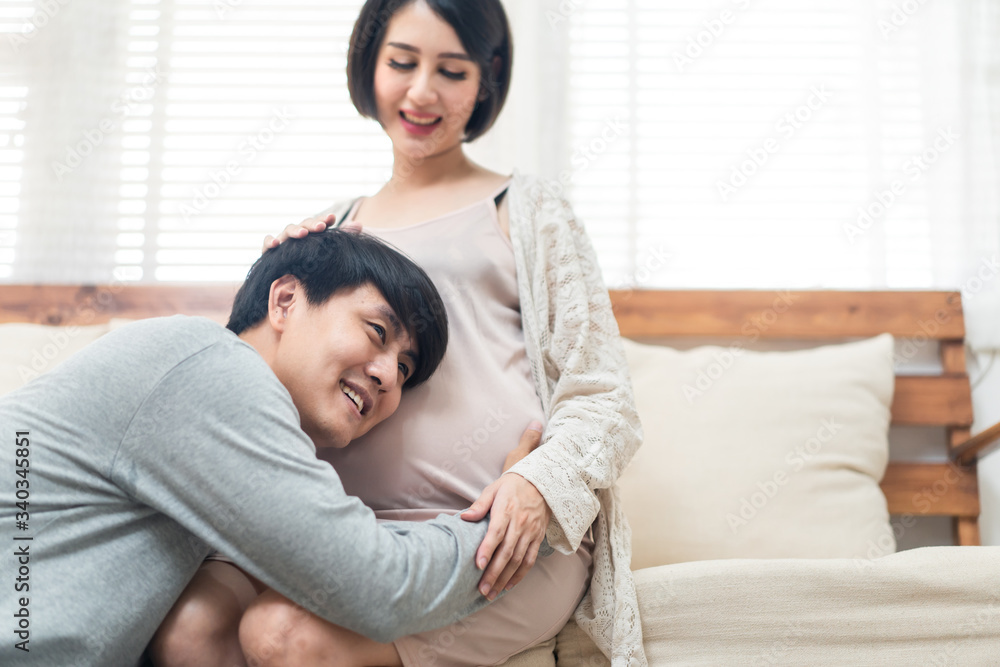 Asian man and pregnancy woman sit on sofa at home. Father holding mother tummy listen and hear infant inside mom belly. Husband sits and hugs wife. Love and care of pregnant and newborn kid concept. Asian man and pregnancy woman sit on sofa at home. Father holding mother tummy listen and hear infant inside mom belly. Husband sits and hugs wife. Love and care of pregnant and newborn kid concept.