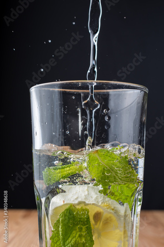 Close-up of glass with mint and lemon with water falling on wooden table and black background, vertical with copy space