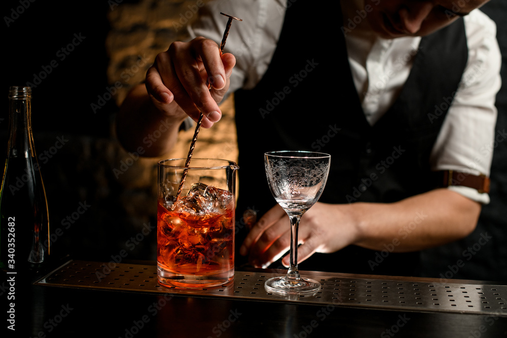Close-up. Bartender's hand holds long spoon and mix drink in glassy mixing cup with pieces of ice Close-up. Bartender's hand holds long spoon and mix drink in glassy mixing cup with pieces of ice