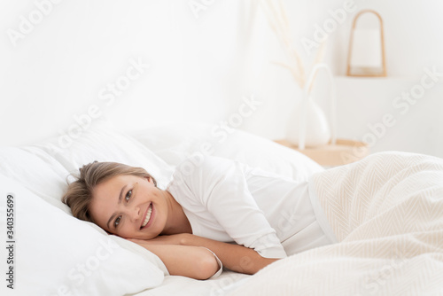 Young female lying in white bed in her bedroom, resting head on palms, looking at camera with positive smile, feeling calm