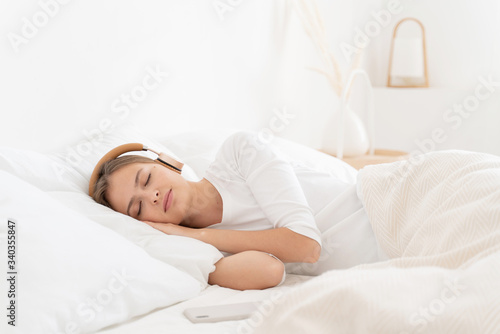 Young woman dressed in white top, lying in bed in morning with eyes closed, listening to calm music via headphones and smartphone