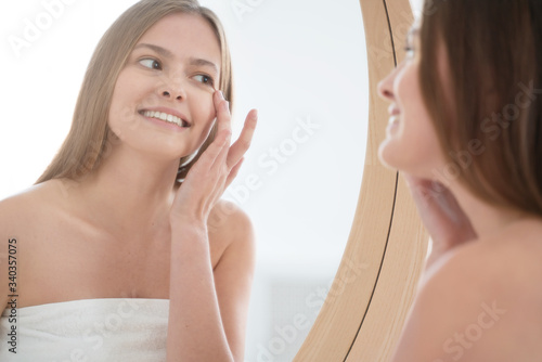 Young woman touching her skin on cheekbone as if applying cosmetic facial treatment, enjoying beauty procedures in bathroom, smiling happily