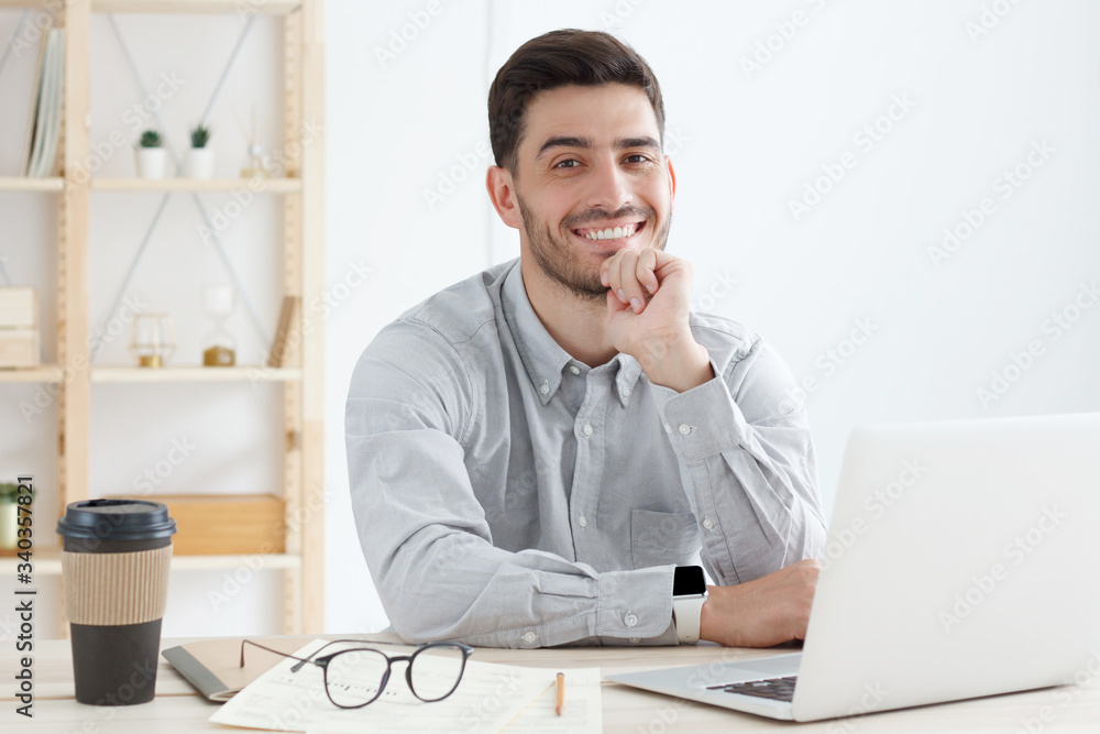 Business guy wearing gray shirt, sitting in front of open laptop, looking at camera with positive smile, ready to help and answer questions Business guy wearing gray shirt, sitting in front of open laptop, looking at camera with positive smile, ready to help and answer questions