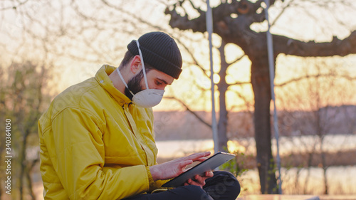 In the middle of the park a guy with a protective mask stay on his tablet