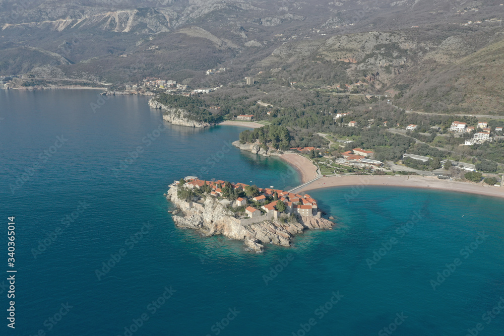 Drone flying over the sea coast, the azure sea and a small island resort, built up with low stone houses with red roofs. Mountains are visible in the background