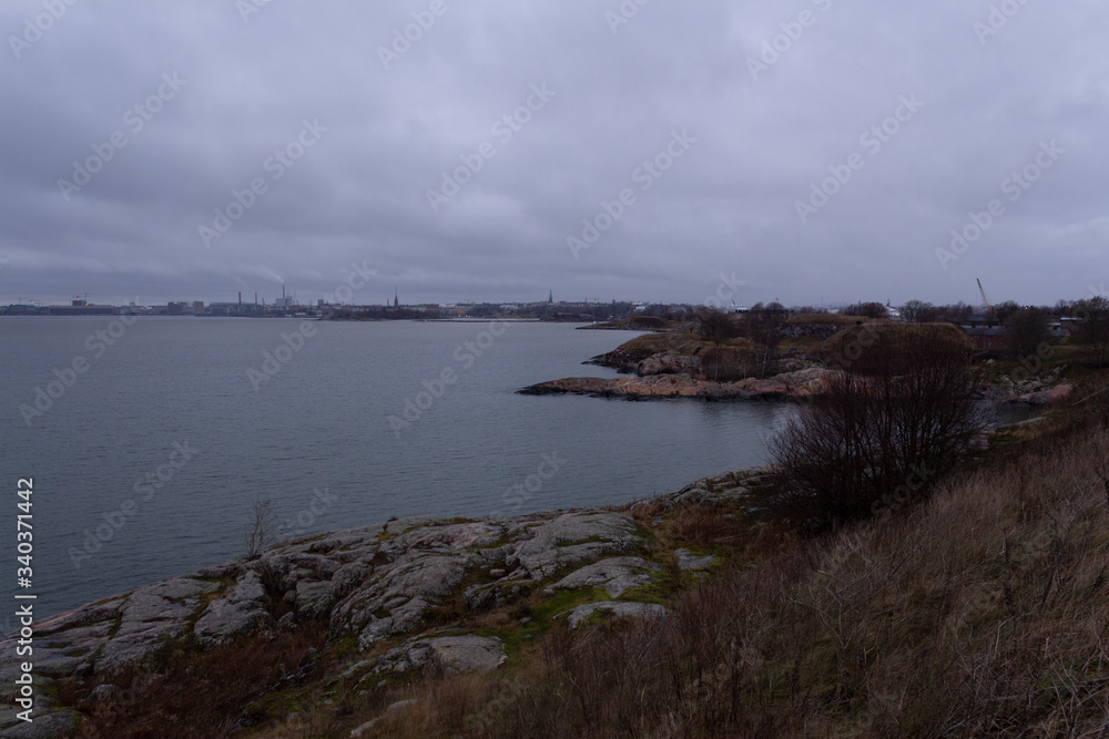 The rocky and grassy shore of Suomenlinna Island and a view of the bay and the city of Helsinki in autumn twilight in Finland. The harsh and laconic beauty of Finnish nature.