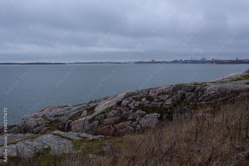 The rocky and grassy shore of Suomenlinna Island and a view of the bay and the city of Helsinki in autumn twilight in Finland. The harsh and laconic beauty of Finnish nature.