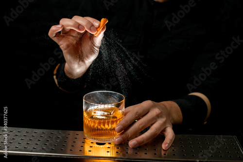 Close-up bartender's hand sprinkle to alcoholic cocktail with piece of citrus zest