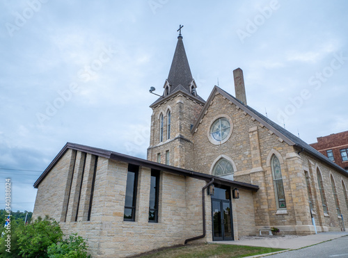 Entrance of a brick Christian Church with a steeple, cross, and stained glass windows on a cloudy evening.