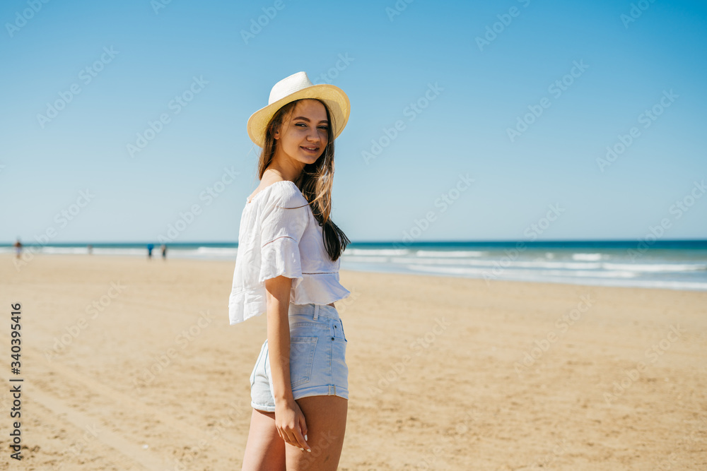slender leggy girl in shorts and a straw hat looks over her shoulder against the background of the ocean and sandy beach