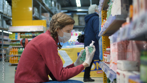 Young woman in a supermarket in a medical mask and rubber gloves chooses a liquid soap for washing hands. Coronavirus pandemic. Precautions against virus infection.