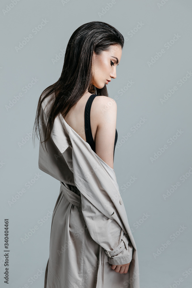Fashion portrait of a young sexy stylish brunette woman in black bodysuit, beige trench coat on a gray background in the studio. Model with perfect skin and professional liner make-up Fashion portrait of a young sexy stylish brunette woman in black bodysuit, beige trench coat on a gray background in the studio. Model with perfect skin and professional liner make-up