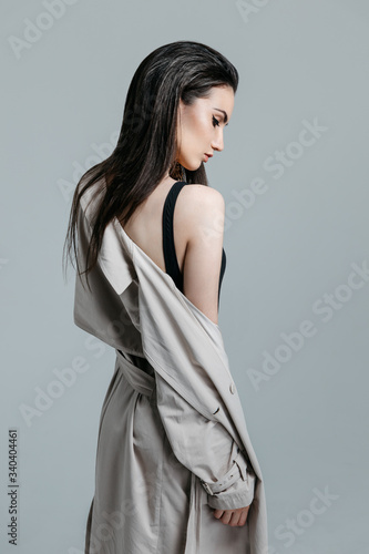 Fashion portrait of a young sexy stylish brunette woman in black bodysuit, beige trench coat on a gray background in the studio. Model with perfect skin and professional liner make-up