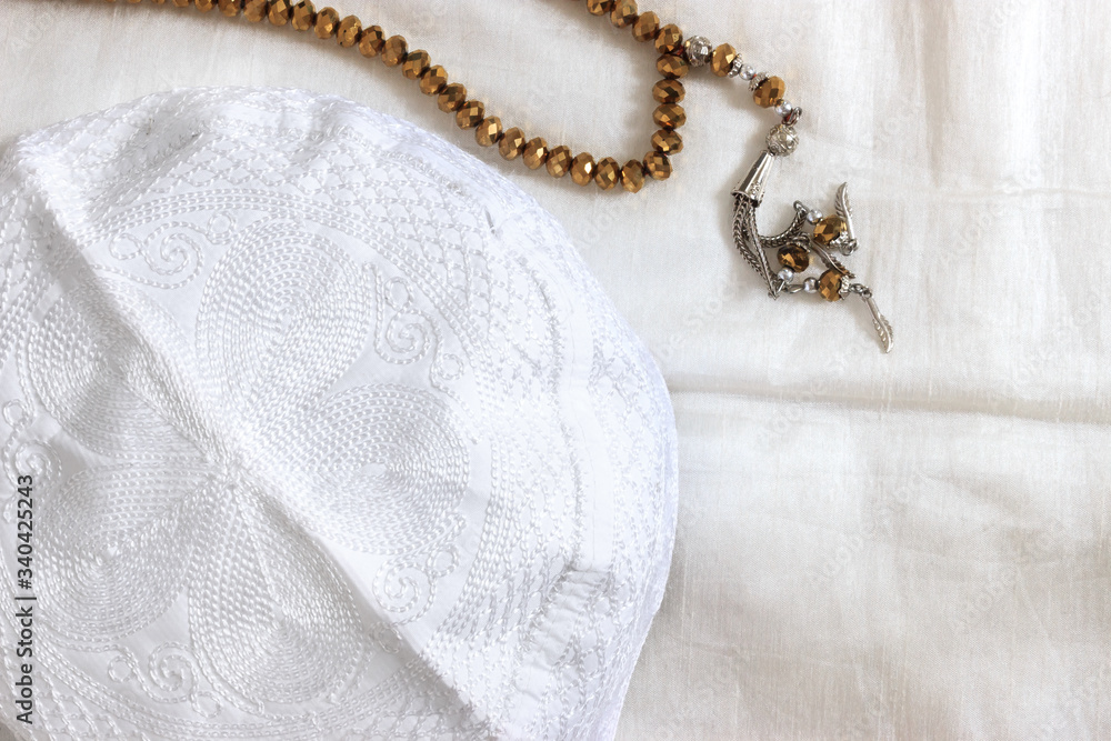 Fototapeta premium Close up top wiew of golden prayer beads and white skullcap on white prayer rug. Muslim pray at home in Ramadan concept.