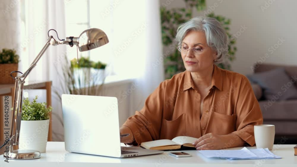 Elderly lady in casual clothes using laptop and making notes then reading document while working on remote business project at home Elderly lady in casual clothes using laptop and making notes then reading document while working on remote business project at home