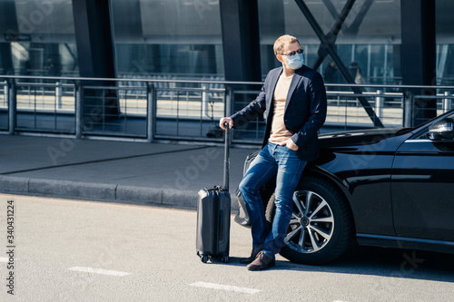 Horizontal full length shot of European man in medical mask looks away, returns from trip, stands with luggage near black car, cares about safety travel, protects himself from coronavirus spread. © VK Studio