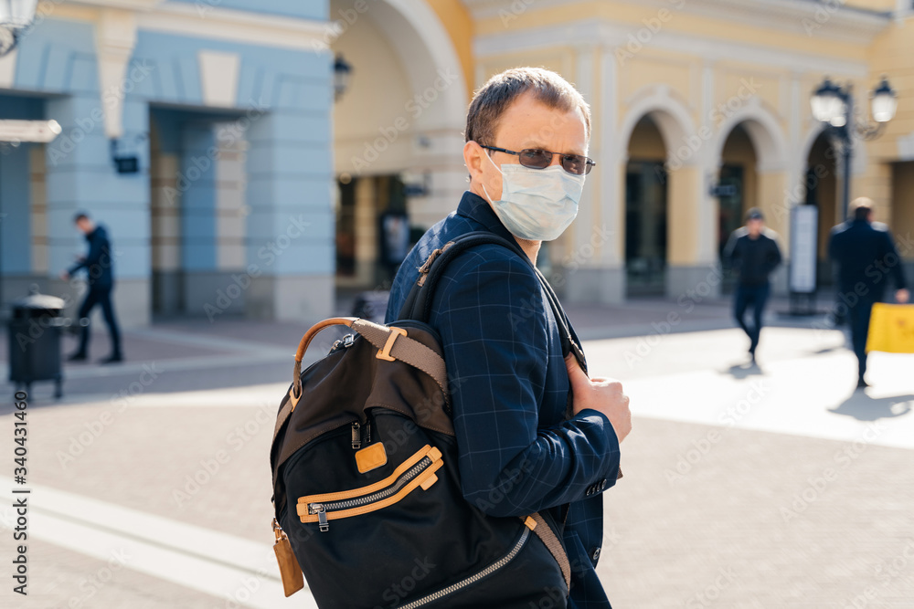 Back view of man traveler wears medical mask for virus prevention carries rucksack on shoulder poses in public place, bus or train station, stands outdoor, proects from coronavirus, contagious disease Back view of man traveler wears medical mask for virus prevention carries rucksack on shoulder poses in public place, bus or train station, stands outdoor, proects from coronavirus, contagious disease