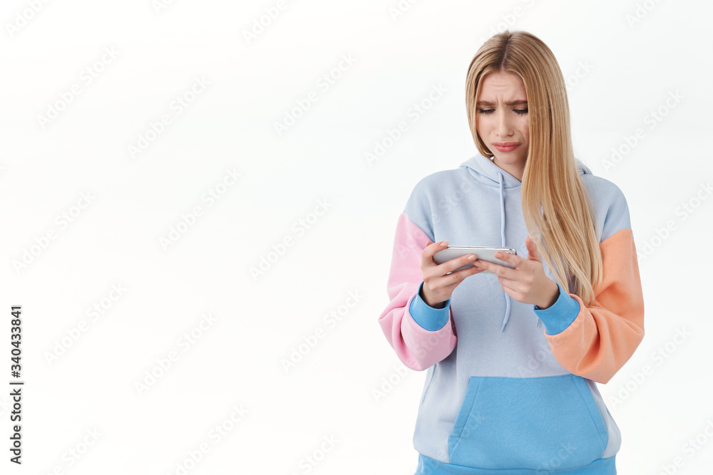 Upset, pensive blonde girl feel uneasy over failure, watching sad movie on mobile phone, sobbing and frowning distressed, holding mobile phone horizontally, standing disappointed white background Upset, pensive blonde girl feel uneasy over failure, watching sad movie on mobile phone, sobbing and frowning distressed, holding mobile phone horizontally, standing disappointed white background