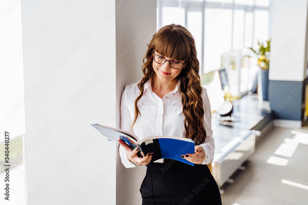 Smart girl with glasses, wearing classic office clothes, stands in coworking space, leaning on the wall. Holding working materials, books, papers and reading them Smart girl with glasses, wearing classic office clothes, stands in coworking space, leaning on the wall. Holding working materials, books, papers and reading them