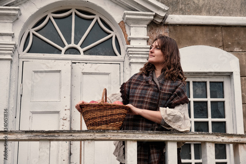 A young woman in vintage clothes and a shawl stands with a basket of apples at the railing of an ancient staircase