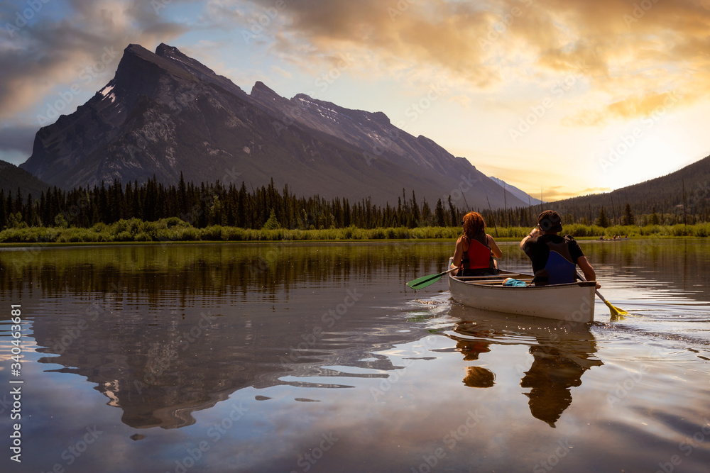Couple adventurous friends are canoeing in a lake surrounded by the Canadian Mountains. Dramatic Sunset Composite. Taken in Vermilion Lakes, Banff, Alberta, Canada. Couple adventurous friends are canoeing in a lake surrounded by the Canadian Mountains. Dramatic Sunset Composite. Taken in Vermilion Lakes, Banff, Alberta, Canada.