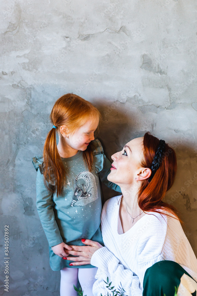 Little girl with red hair, braided ponytails, in a dress, stands near the wall and hugs mom, communicating with a parent, relationships, family, attention, emotions, feelings, significance, upbringing